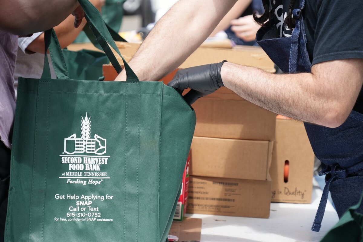 Two people pack food into a green Second Harvest Food Bank tote bag at a food distribution event, offering neighbor assistance as cardboard boxes are stacked in the background.