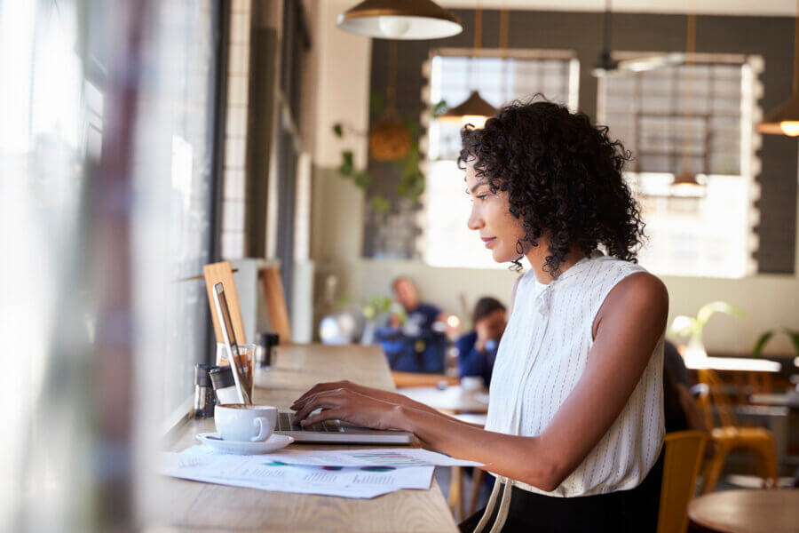 The Money Moves That Matter Most for Women (in Every Decade!) - 3 Woman working on a laptop at a cafΓ© counter, with documents and a cup of coffee in front of her, focused on financial planning for women. Other people are visible in the background.
