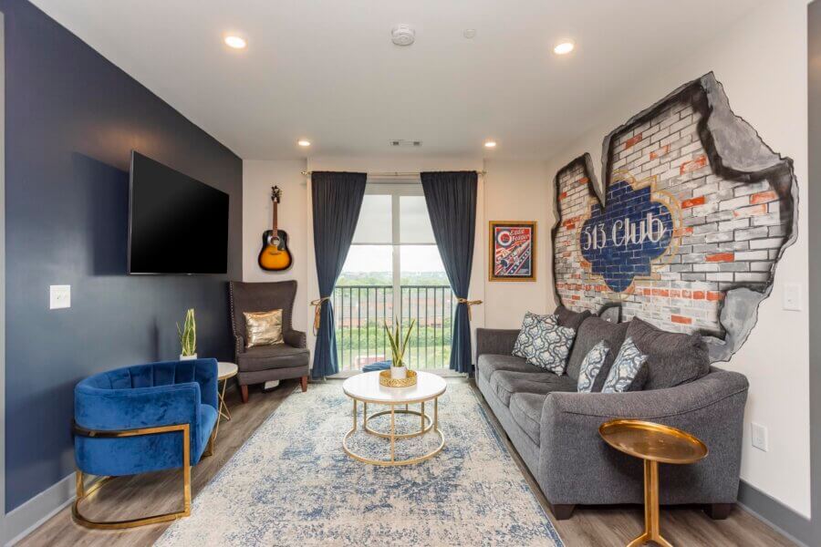 Modern living room at 65 Lindsley Ave, Nashville, TN, featuring a gray sofa, blue accent chair, wall art, mounted guitar, round coffee table, and floor-to-ceiling window with curtains.