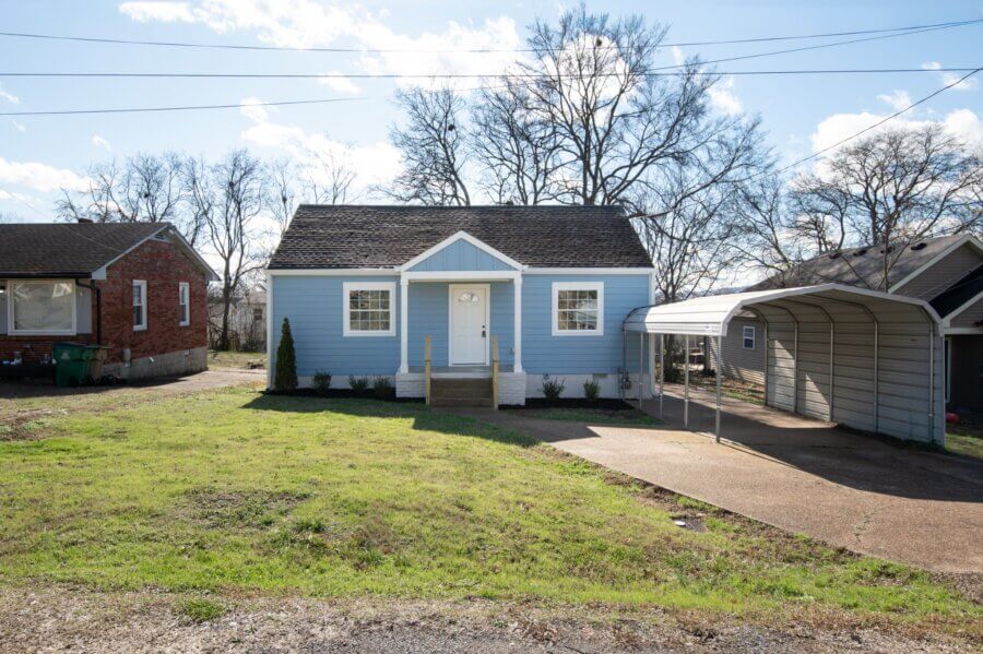 A small, light blue house with a front porch, white trim, sloped roof, and attached carport on the right sits on a grassy lot at 112 Wiley St, Madison TN, with leafless trees providing a charming backdrop.