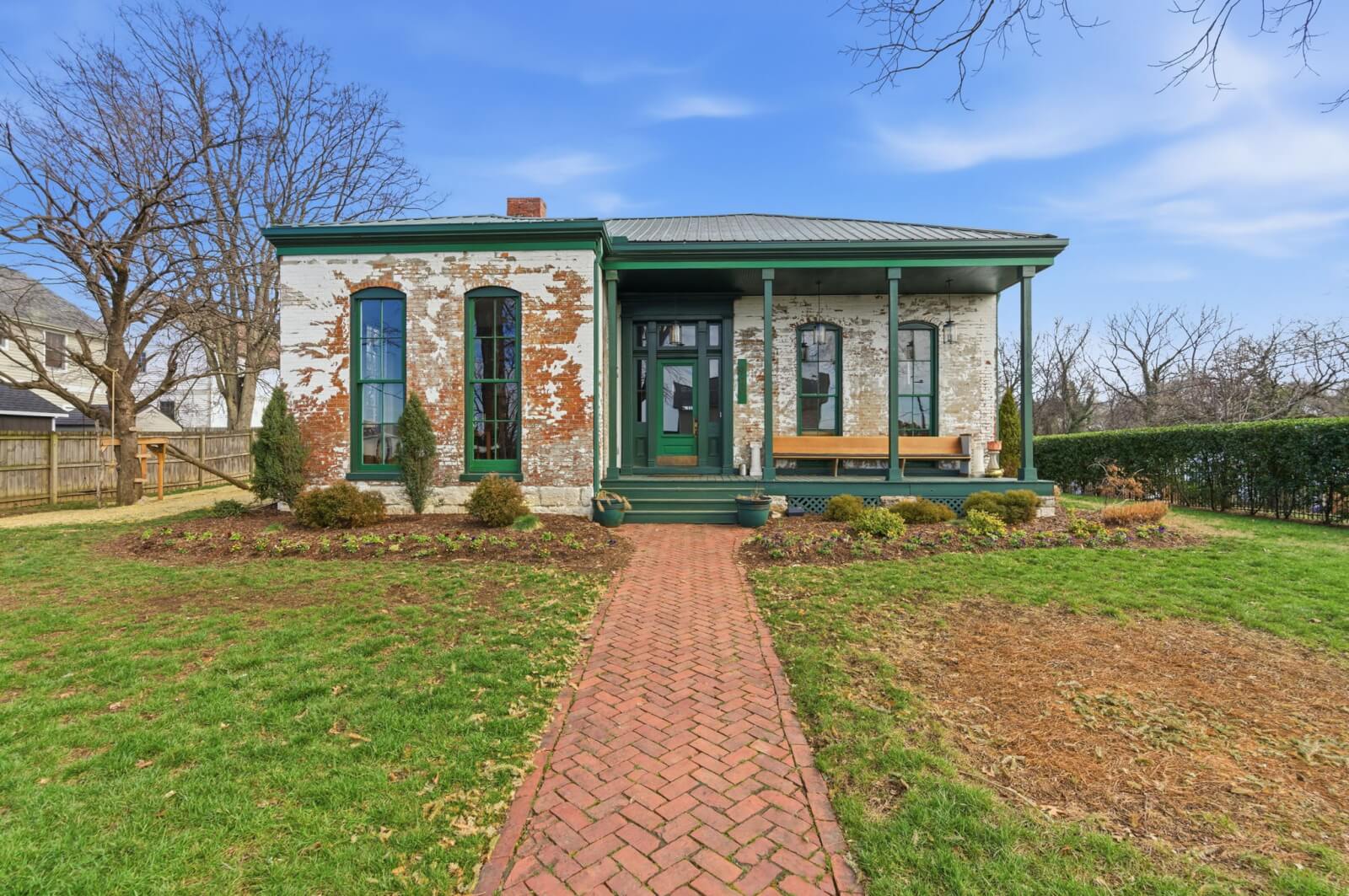 Charming single-story brick house in Nashville with weathered white paint, green trim, a covered front porch, and a red brick walkway at 909 Manila Ave—surrounded by grass and trees under a blue sky.