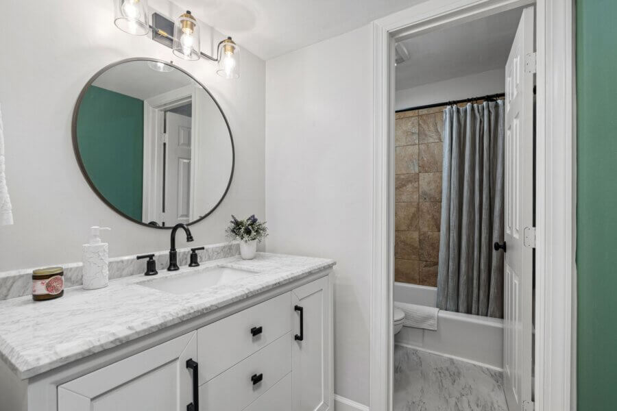 Modern bathroom at 4425 Westlawn Dr, Nashville, TN features a marble countertop vanity, round mirror, and black fixtures; adjacent room offers a bathtub with tan tile and a gray shower curtain.