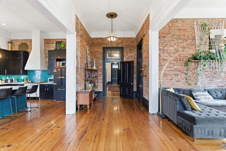 Open-concept living space at 909 Manila Ave features exposed brick walls, wood floors, a modern kitchen with bar stools, and a gray sectional sofa accented by wall plants—a stylish Nashville retreat.