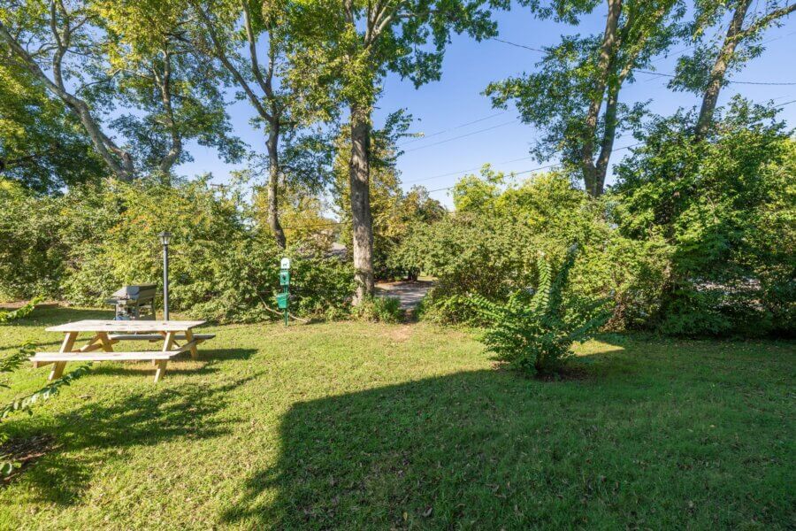 A grassy backyard at 4425 Westlawn Dr, Nashville TN, features a wooden picnic table, grill, and lamp post, surrounded by trees and shrubs under a clear blue sky.
