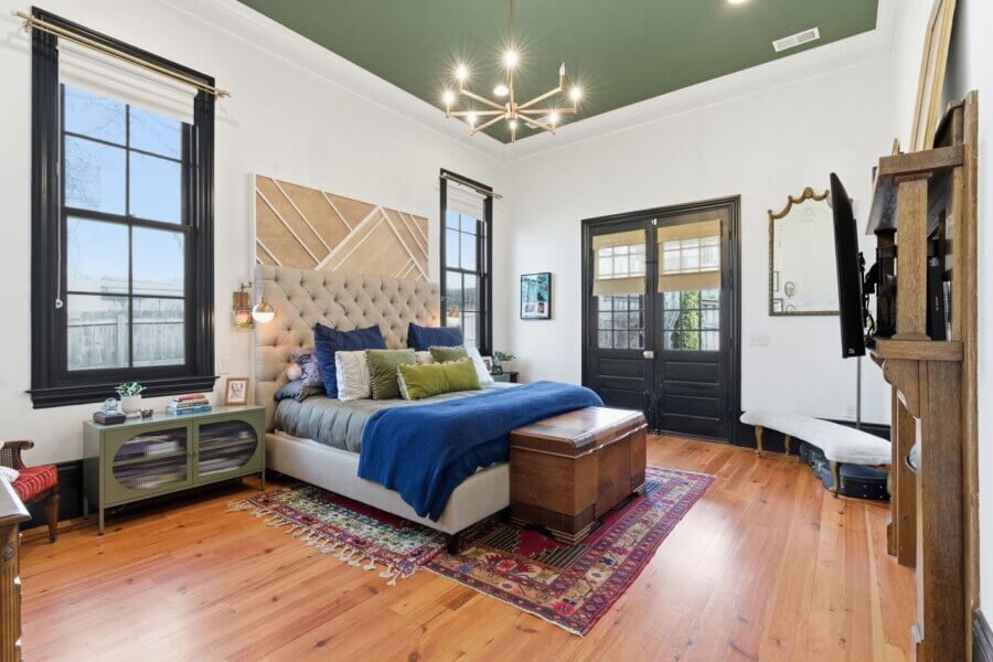 A modern bedroom in Nashville TN with a green ceiling, wooden floors, a large bed with blue bedding, a chandelier, two windows, a glass door, and a colorful rug under the bed.