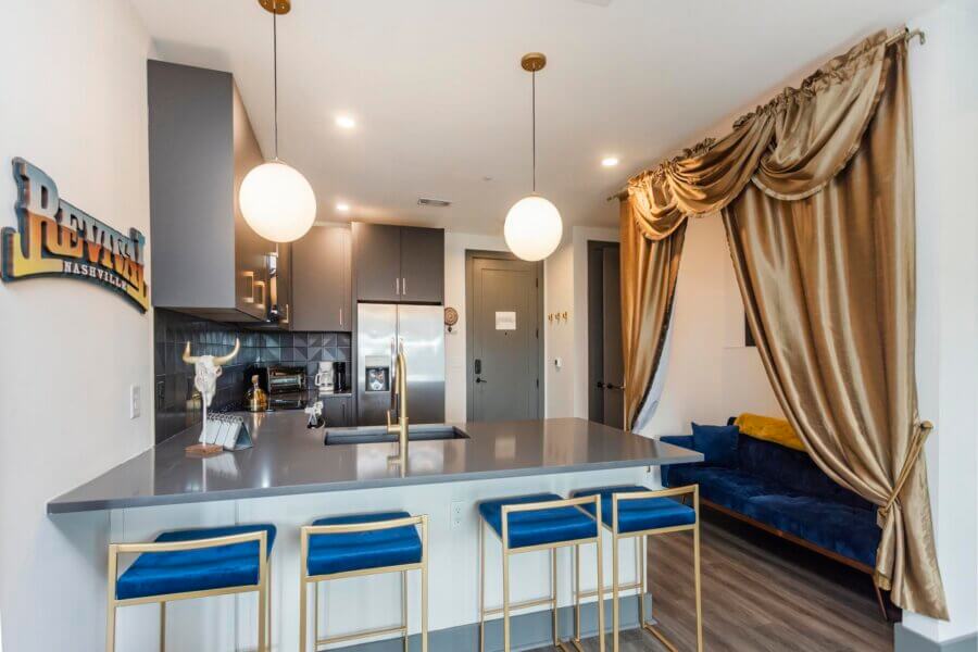 Modern kitchen at 65 Lindsley Ave, Nashville TN, with gray cabinets, stainless steel appliances, and a breakfast bar with blue stools; gold curtains frame a blue sofa in the adjacent living area.