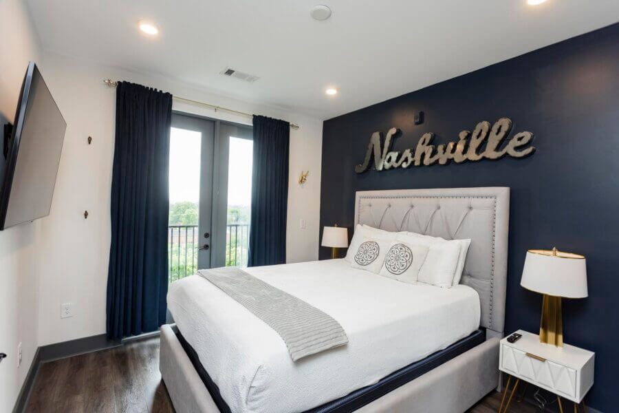 A modern bedroom with a gray upholstered bed, white bedding, bedside tables with lamps, dark curtains, and a sign reading "Nashville" on a navy accent wall brings 65 Lindsley Ave's charm to life.