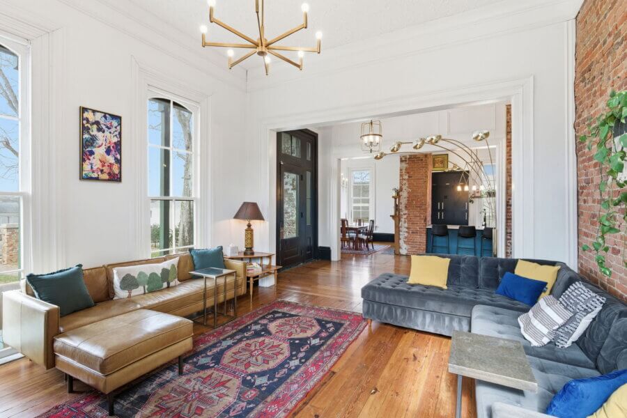 Bright living room at 909 Manila Ave, Nashville TN, featuring large windows, a brown leather sofa, gray sectional, colorful rugs, wood floors, exposed brick wall, and modern chandeliers. Dining area visible in the background.