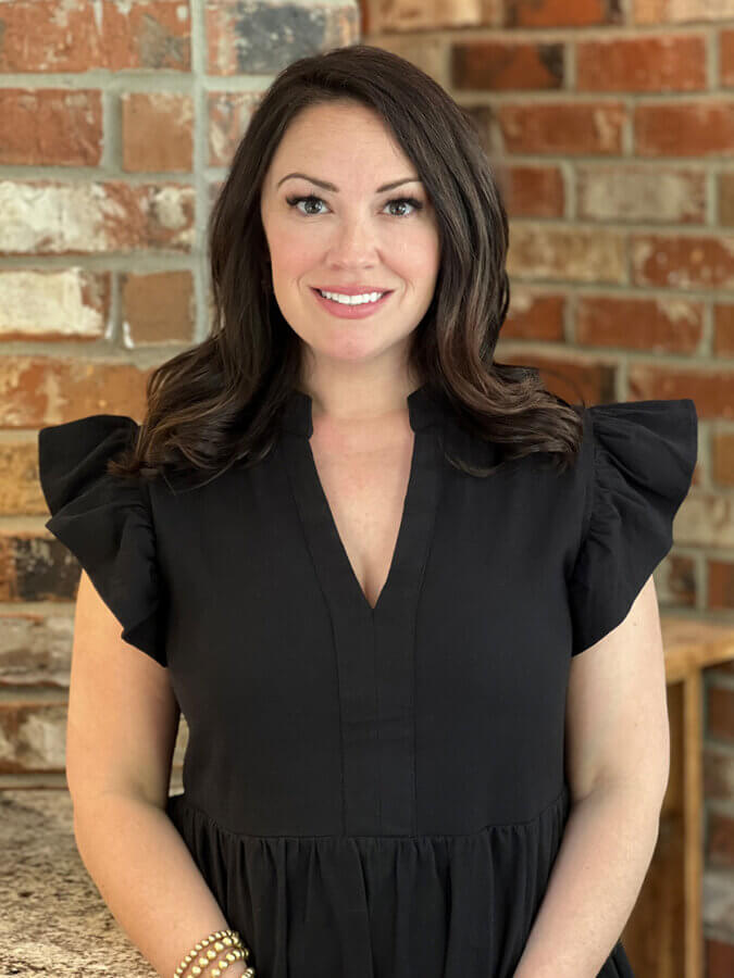 A woman with dark hair wearing a black dress stands in front of a brick wall, smiling at the camera on Westlawn Dr in Nashville.