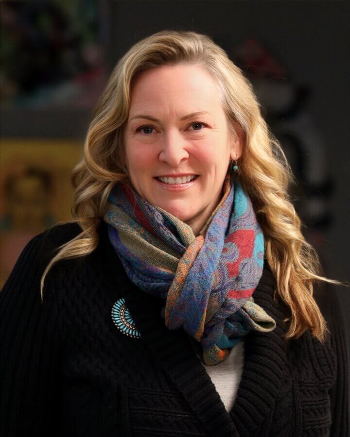 She's Building a Support Network for Nashville's Live Music Community - 6 Kara Kemp, with wavy blonde hair, smiles at the camera, wearing a patterned scarf, black sweater, and a turquoise brooch, set against a blurred indoor background.