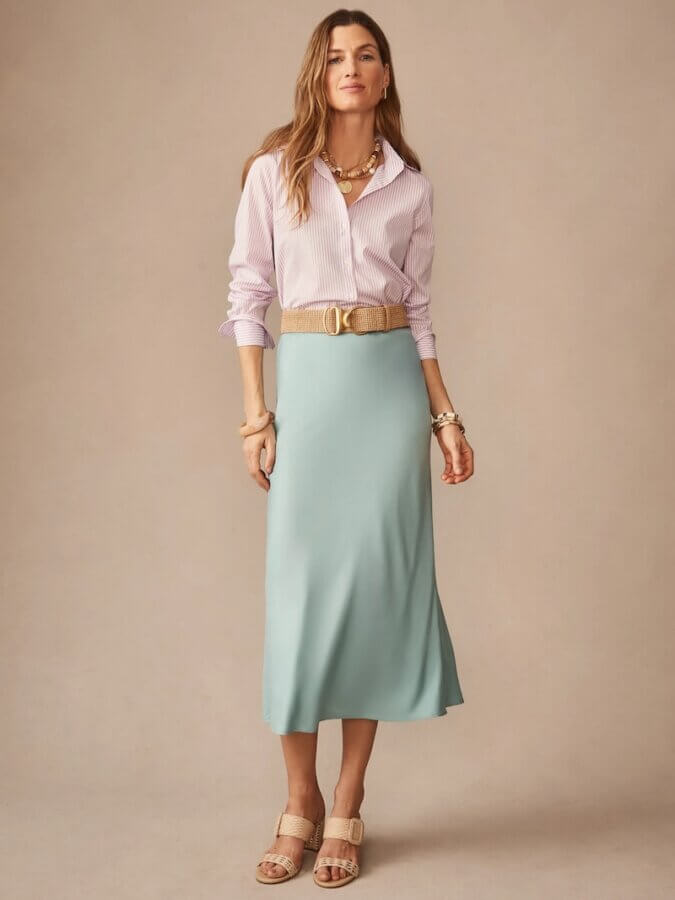 6 Spring 2026 Fashion Trends + How to Wear Them - 11 A woman stands wearing a light pink striped shirt, a light blue midi skirt, a beige belt, beige sandals, and gold jewelry against a plain beige background—an effortless take on How to Wear Spring 2026 Fashion Trends.