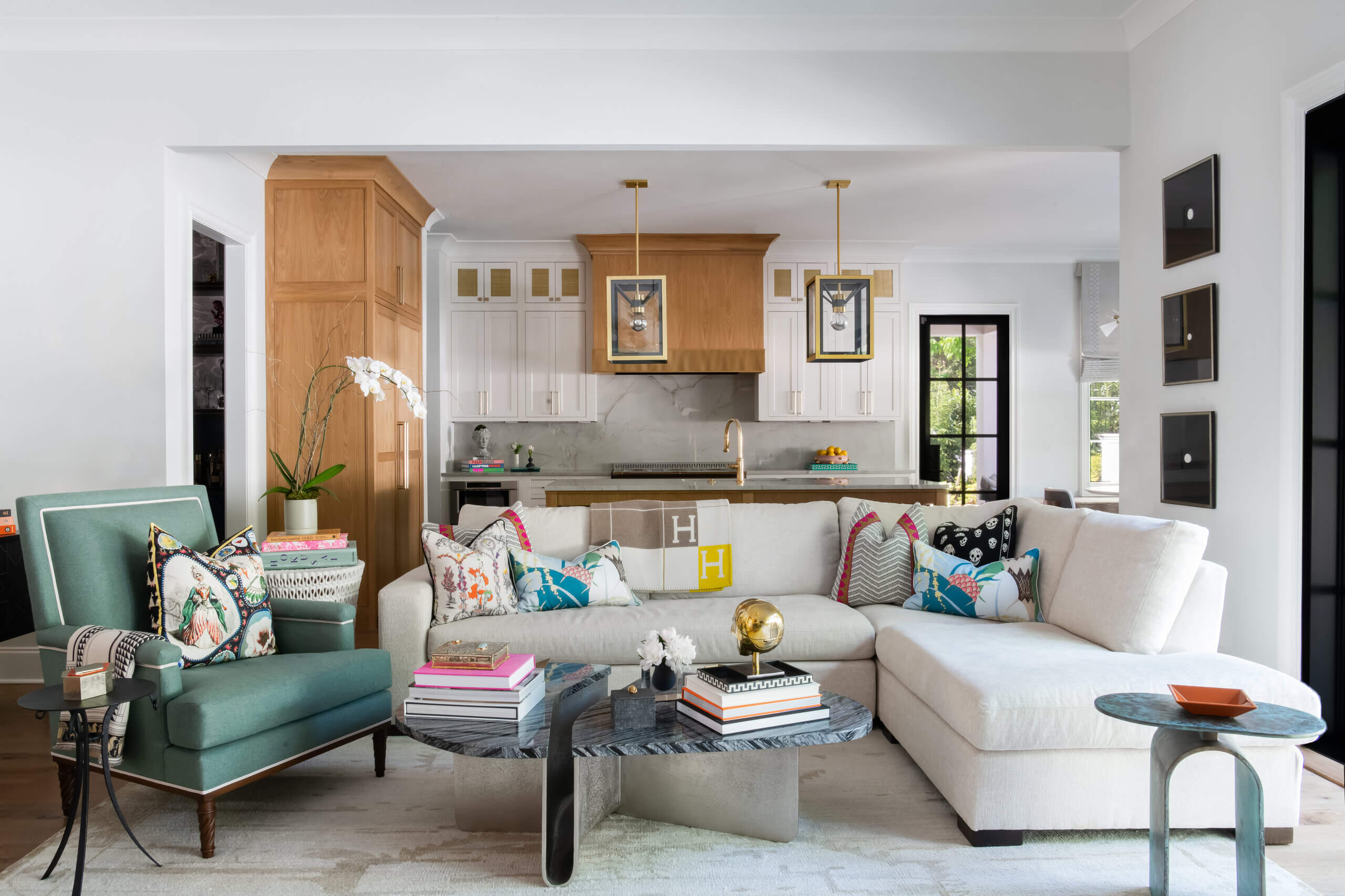 Modern living room designed by JASPER Builds, featuring a white sectional sofa, colorful pillows, green armchair, marble coffee table, and an open kitchen with wood accents in the background.