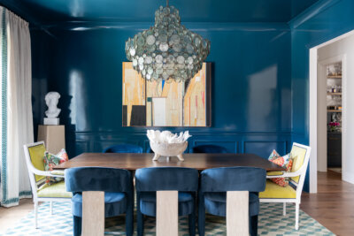 A JASPER Builds dining room with glossy blue walls, a wooden table, blue and yellow chairs, large round chandelier, geometric rug, wall art, and a sculpture on a pedestal.
