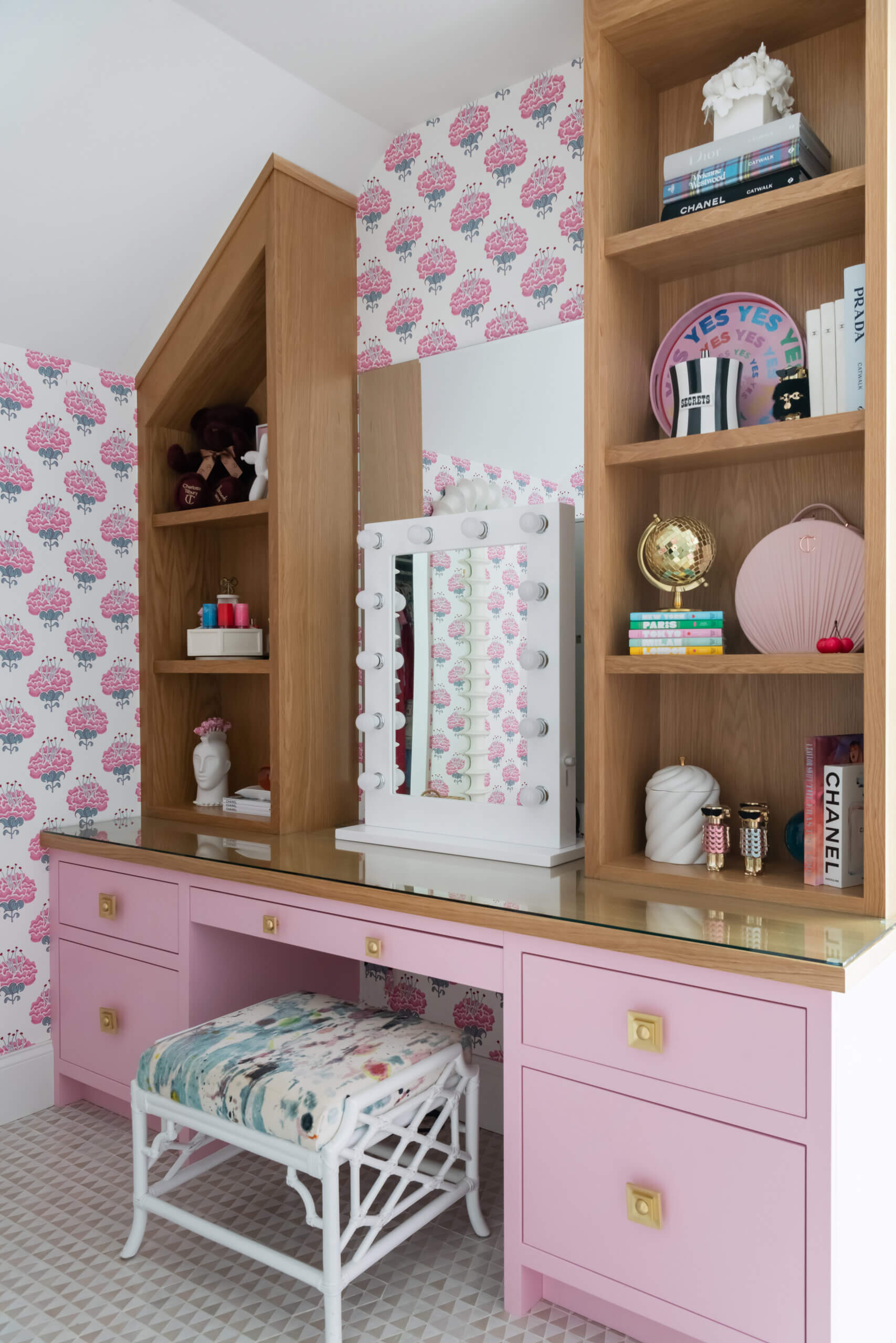 A JASPER vanity with pink drawers, a built-in mirror with bulbs, wood shelving holding books and decor, and a patterned stool, set against pink floral wallpaper—beautifully blends stylish Builds and function.
