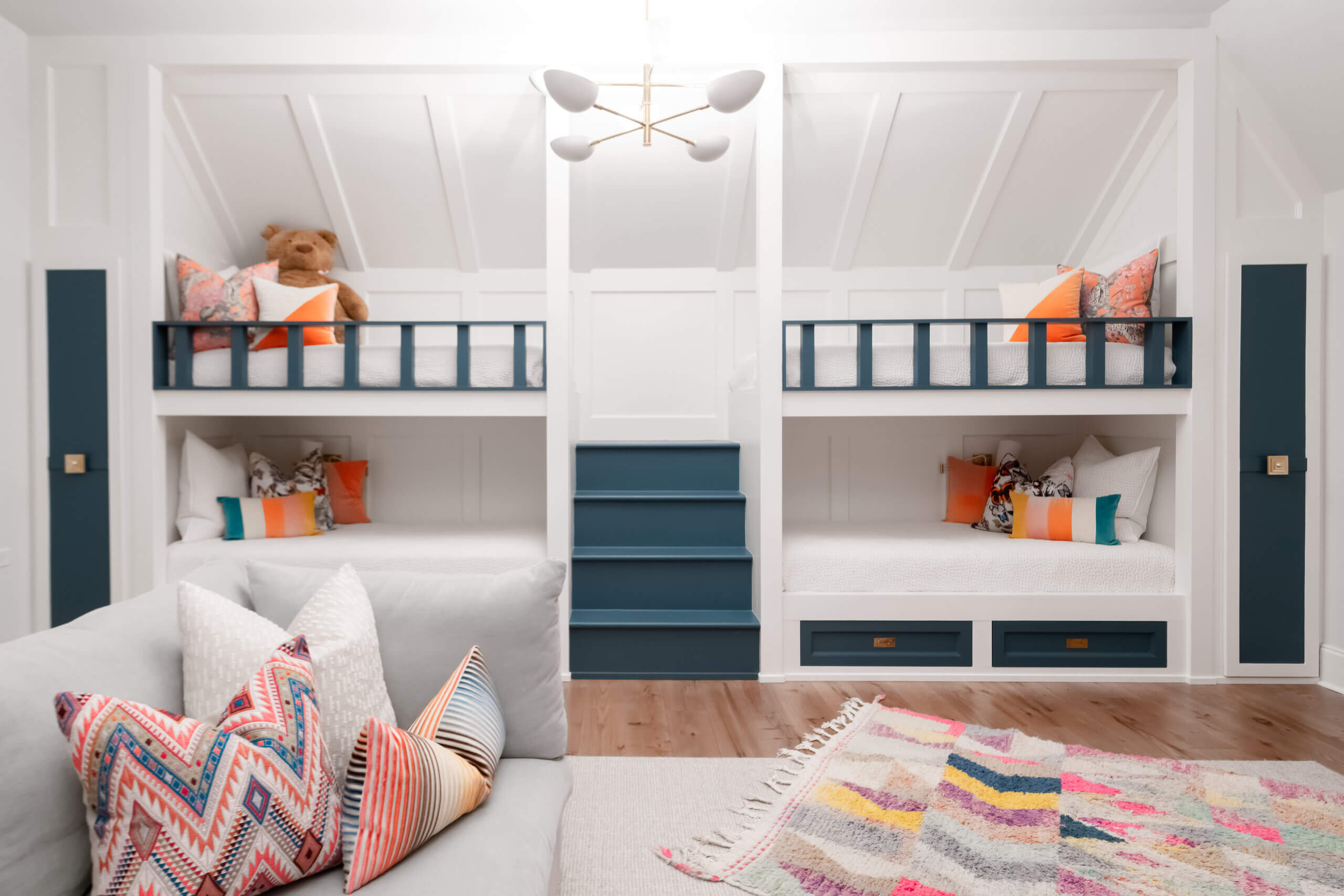 A bright, modern bedroom by JASPER Builds features two sets of built-in bunk beds, colorful pillows, a large teddy bear, a patterned rug, and a gray sofa in the foreground.