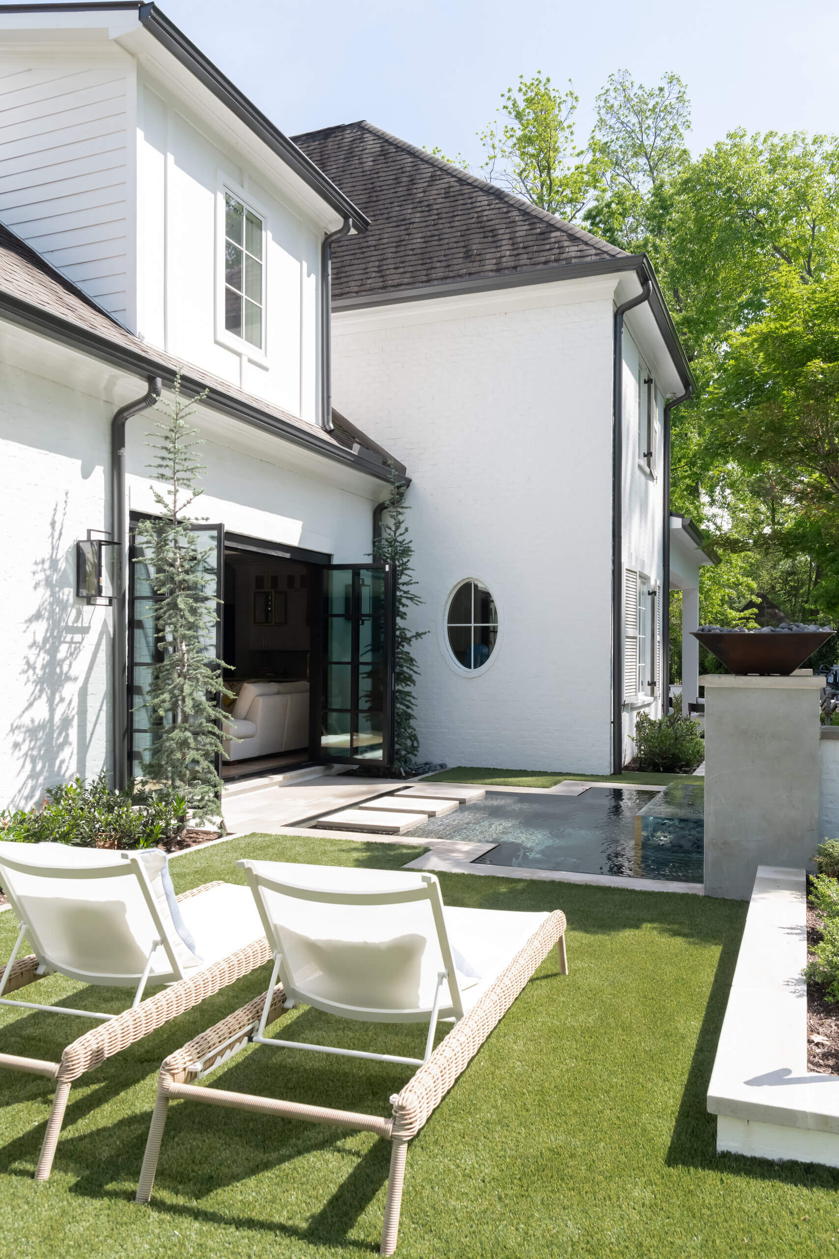Two lounge chairs face a small rectangular pool in a modern backyard with artificial grass, white exterior walls, and large glass doors—showcasing the signature style of JASPER Builds.