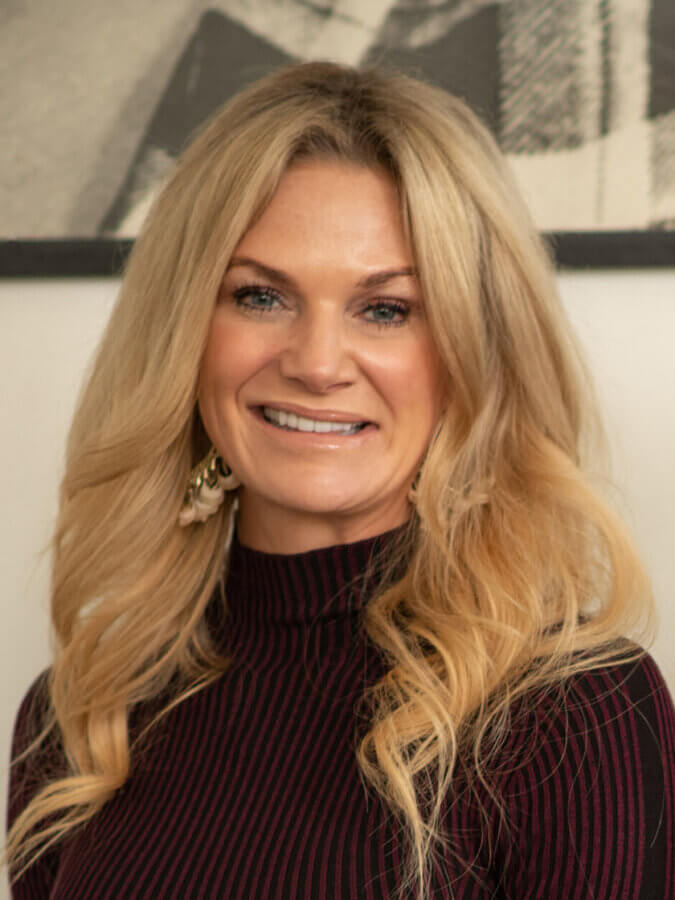 A woman with long blonde hair wearing a dark, high-neck top, smiling at the camera with a blurred black-and-white photo in the background at 4425 Westlawn Dr, Nashville.