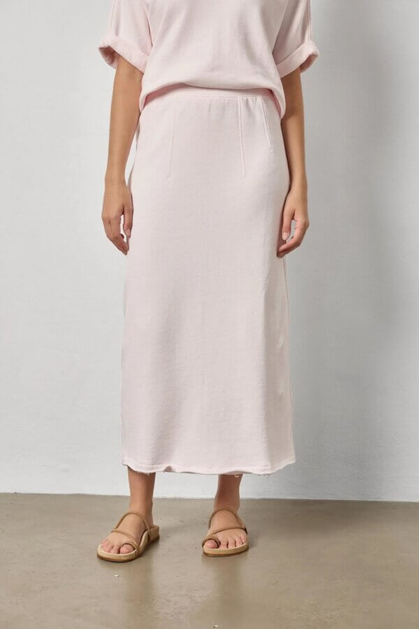 6 Spring 2026 Fashion Trends + How to Wear Them - 18 A person wearing a light pink midi skirt and matching top stands against a plain white wall, with tan sandals on a concrete floor—showcasing effortless style inspired by Spring 2026 Fashion Trends.