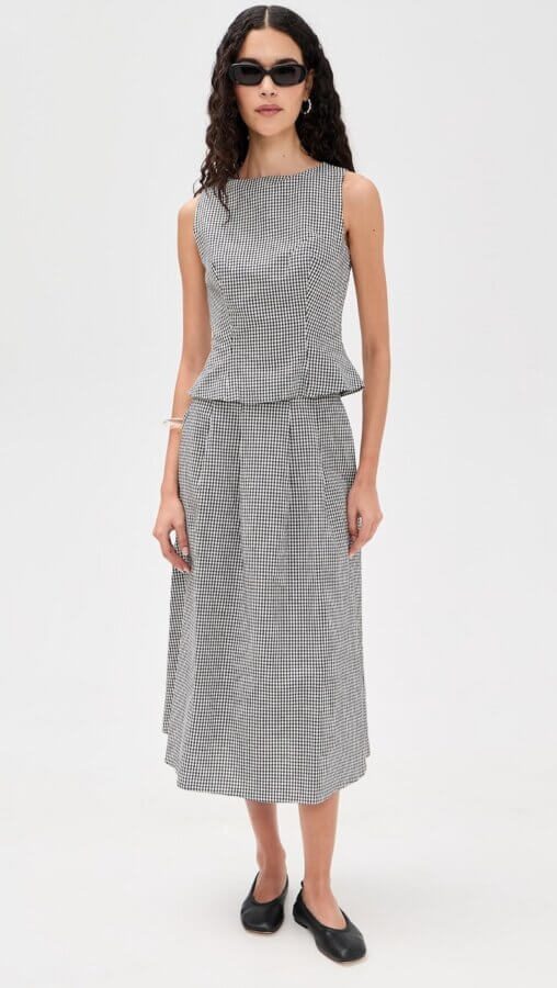 6 Spring 2026 Fashion Trends + How to Wear Them - 16 A woman embraces Spring 2026 Fashion Trends in black sunglasses, a sleeveless black and white checkered top, and a matching midi skirt with black flats, standing against a plain, light background.