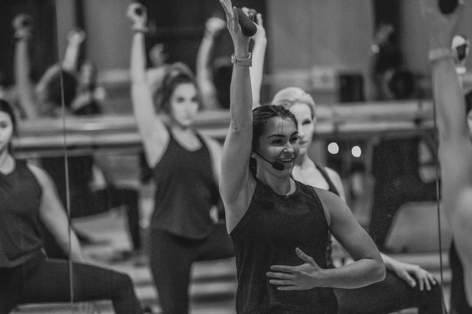A group fitness instructor leads an exercise class using the PM Method, raising one arm as participants follow along in a mirrored studio.