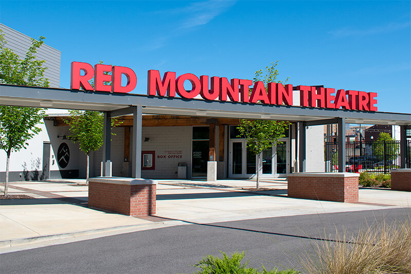 The exterior entrance of Red Mountain Theatre features bold red signage and a covered walkway, showcasing the vibrant spirit of Red Mountain Theatre on a clear day.