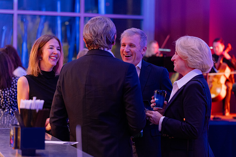 Four people in formal attire stand together, smiling and conversing at an indoor Red Mountain Theatre event with musicians performing in the background.