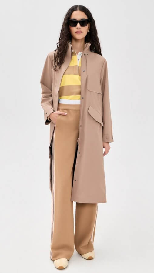 6 Spring 2026 Fashion Trends + How to Wear Them - 2 A person wearing sunglasses, a beige long coat, a yellow and white striped shirt, wide-leg tan pants with stripes, and beige shoes stands against a plain light background.
