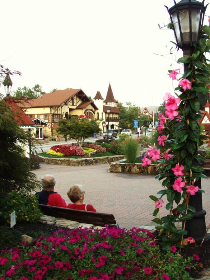 9 Hidden Gems to Visit in the South - 8 Two people sit on a bench in a flower-filled town square, a hidden gem in the South, with Alpine-style buildings and a streetlamp adorned with pink flowers in the foreground.