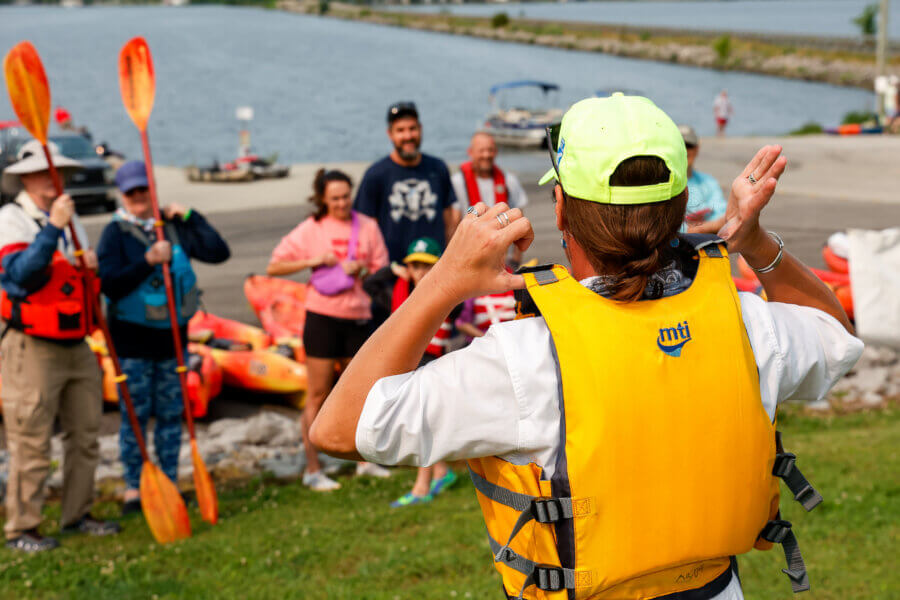 52 Weekends, 630 Miles: Alabama’s Scenic River Trail is the Longest in the Country - 9 A person in a yellow life jacket addresses a group holding paddles near kayaks by the waterfront, inviting them to explore Alabama on weekends on the water along the scenic Alabama River Trail.