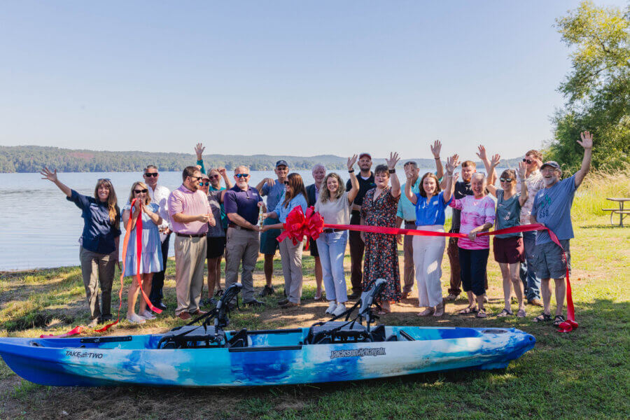 52 Weekends, 630 Miles: Alabama’s Scenic River Trail is the Longest in the Country - 8 A group of people stand by a lake, smiling and waving, at a ribbon-cutting ceremony with a blue tandem kayak in the foreground—ready to explore Alabama along the Alabama Scenic River Trail and enjoy weekends on the water.