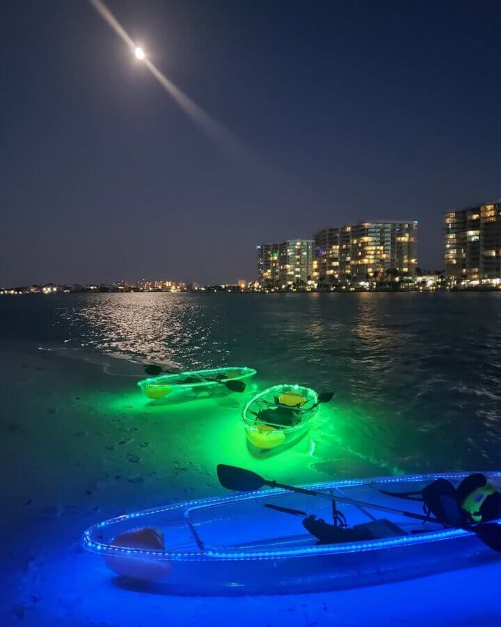 52 Weekends, 630 Miles: Alabama’s Scenic River Trail is the Longest in the Country - 3 Three illuminated kayaks sit on a beach at night under a full moon, city lights twinkling across the water.