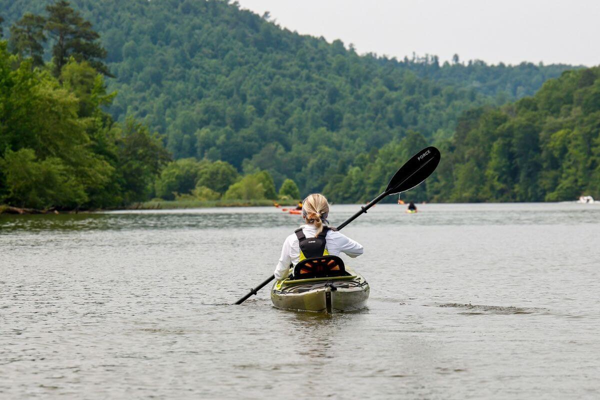 A person paddles a kayak on a calm river along the Alabama Scenic River Trail, surrounded by green, forested hills under an overcast sky—perfect for weekends on the water.