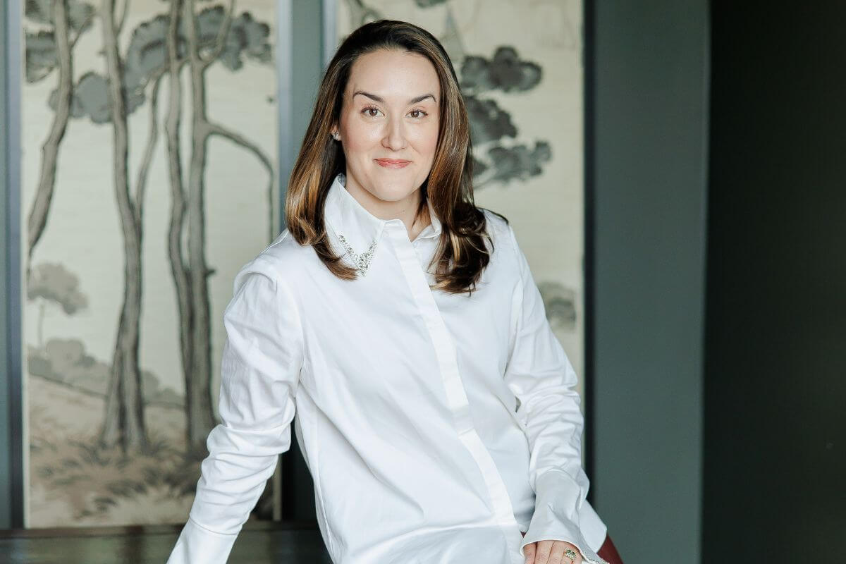 Ellie Christopher, an interior designer with long brown hair, stands indoors in front of a wall adorned with tree artwork, wearing a white button-down shirt.