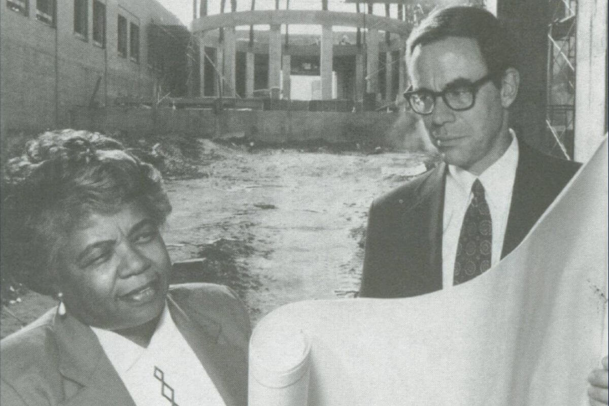 Two women in business attire stand at a Birmingham construction site, examining a large rolled document. An unfinished building structure is visible in the background, highlighting women's roles in shaping the city's future.