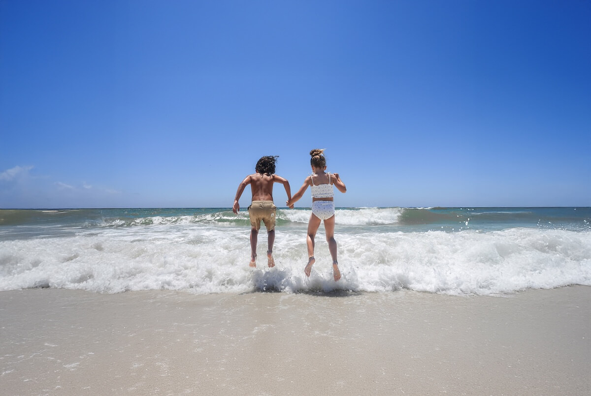 Two children holding hands jump into the ocean waves on a fabulous beach, with clear blue sky and white surf in the background, capturing a perfect sun-soaked escape.