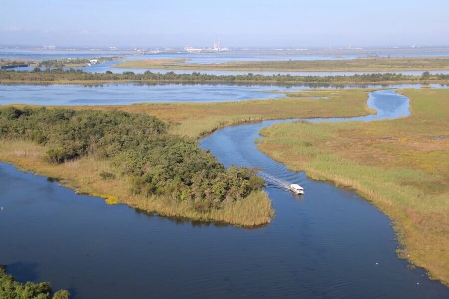52 Weekends, 630 Miles: Alabama’s Scenic River Trail is the Longest in the Country - 6 A small boat travels along the Mobile-Tensaw Delta, surrounded by dense vegetation and wetlands, with industrial buildings visible in the distant background.
