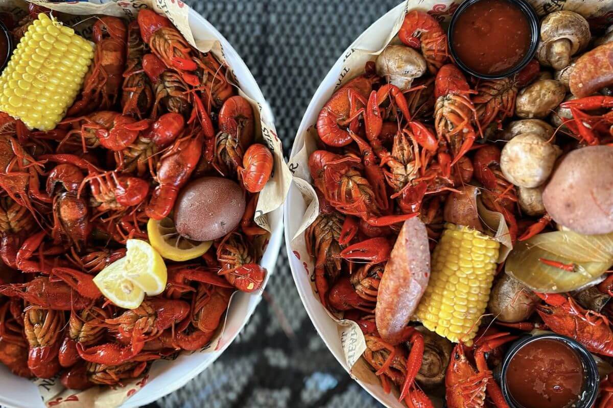 Two bowls filled with boiled Crawfish, corn on the cob, potatoes, sausage, mushrooms, lemon slices, and containers of dipping sauce—a true Birmingham seafood feast guide.