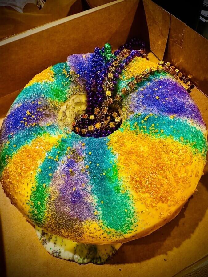 10 New Birmingham Restaurants to Try This Month - 19 A round King Cake decorated with yellow, green, and purple sugar, topped with Mardi Gras beads, sits inside a cardboard box—a festive treat often featured at new restaurants Birmingham has to offer.