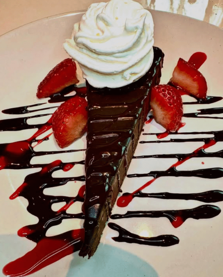 10 New Birmingham Restaurants to Try This Month - 15 A slice of chocolate cake topped with whipped cream, garnished with sliced strawberries and drizzled with chocolate and berry sauces on a white plate.
