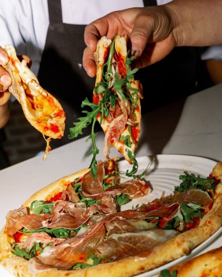 10 New Birmingham Restaurants to Try This Month - 16 A person lifts a slice of pizza topped with arugula and prosciutto from a plate, while another hand holds a folded slice.