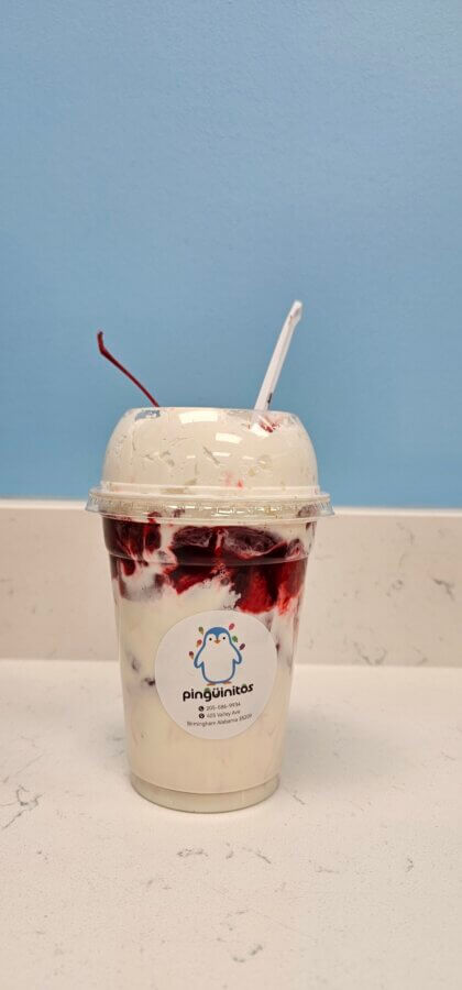 10 New Birmingham Restaurants to Try This Month - 18 A clear plastic cup of vanilla ice cream with cherry topping and a cherry on top, labeled "Pingüinitos," sits on a white counter against a blue wall—a sweet find at one of the new restaurants Birmingham has to offer.
