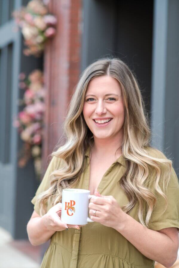 Meet Jessica Watts of BHAM's House Plant Collective - 8 Jessica Watts with long, wavy blonde hair wearing an olive green shirt stands outside holding a white mug with “HPC” printed on it, smiling at the camera.