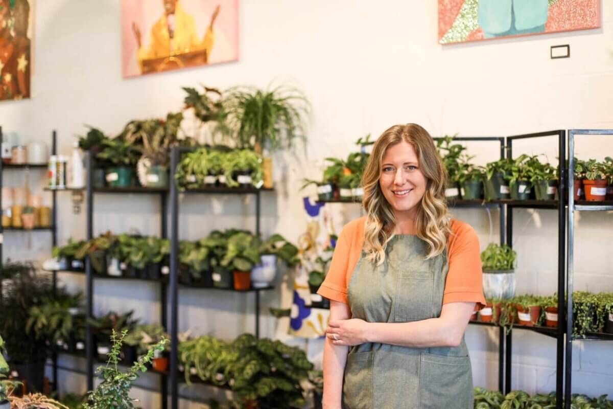 Jessica Watts, wearing an orange shirt and apron, stands in front of shelves filled with potted plants at House Plant Collective’s brightly lit plant store. Photo by Libby Malcom Photography
