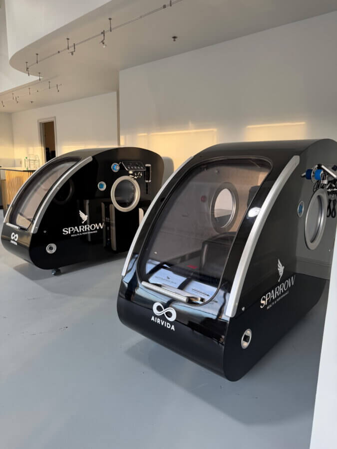 Her BHAM Clinic Blends Holistic Healing with Medical Expertise - 4 Two black hyperbaric oxygen therapy chambers labeled "SPARROW" and "AIRVIDA" stand side by side in a sleek, modern room at Sparrow Clinic.