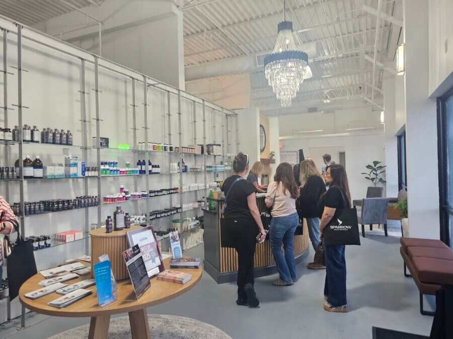 Her BHAM Clinic Blends Holistic Healing with Medical Expertise - 3 People stand in line at a modern wellness or pharmacy store, where shelves of products and informational brochures on a round table reflect an environment bridging the gap between holistic medicine and conventional medicine, beneath a chandelierโs glow.
