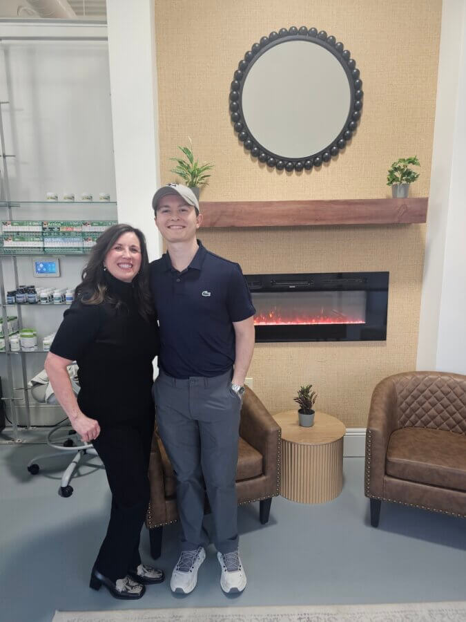 Her BHAM Clinic Blends Holistic Healing with Medical Expertise - 2 Two people stand smiling in front of a modern fireplace, bridging the gap between conventional medicine and holistic medicine, with armchairs, a small plant, and shelves stocked with products in the background.
