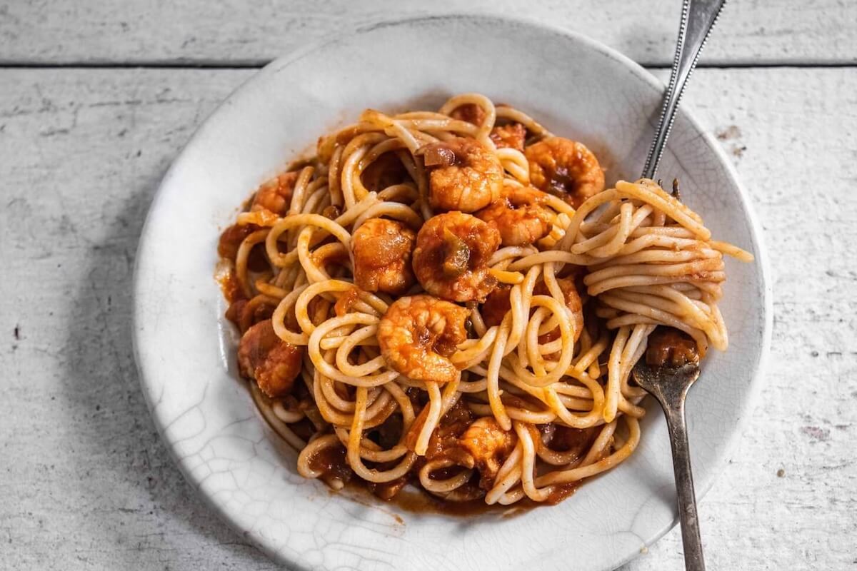 A white plate of spaghetti with shrimp in tomato sauce, with a fork twirling some pasta, sits on a rustic white wooden surface.