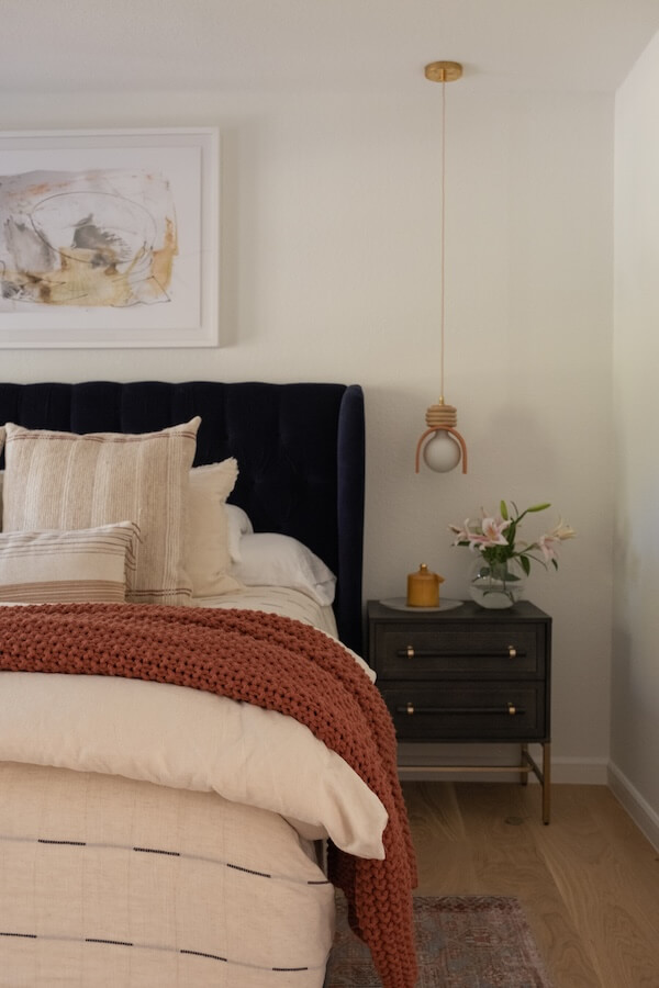 Step Inside an Austin Interior Designer’s Home - 10 A neatly made bed with beige bedding and a rust knit blanket creates a personal retreat beside a dark nightstand with a candle, flowers, and a modern pendant light above. Abstract art hangs on the wall, showcasing thoughtful interior design.