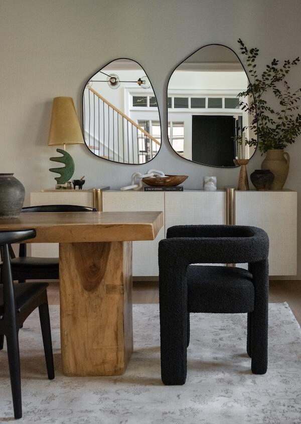Step Inside an Austin Interior Designer’s Home - 15 A modern dining area designed by an Austin interior designer features a wooden table, black chair, sideboard, two irregular mirrors, a lamp, vases, and green plants.