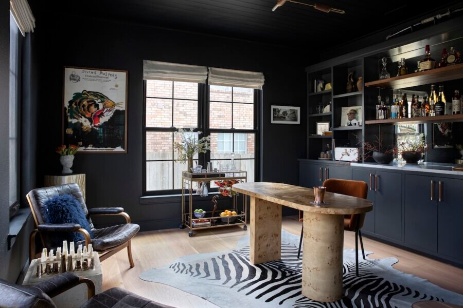 Step Inside an Austin Interior Designer’s Home - 13 A modern home office designed by an Austin interior designer, with dark walls, wooden desk, leather chairs, zebra-patterned rug, wall shelves with bottles, art on the wall, and large windows.